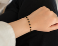 A delicate gold chain bracelet with small black beads worn on a person's wrist, shown against a dark black top and light beige skin tone.