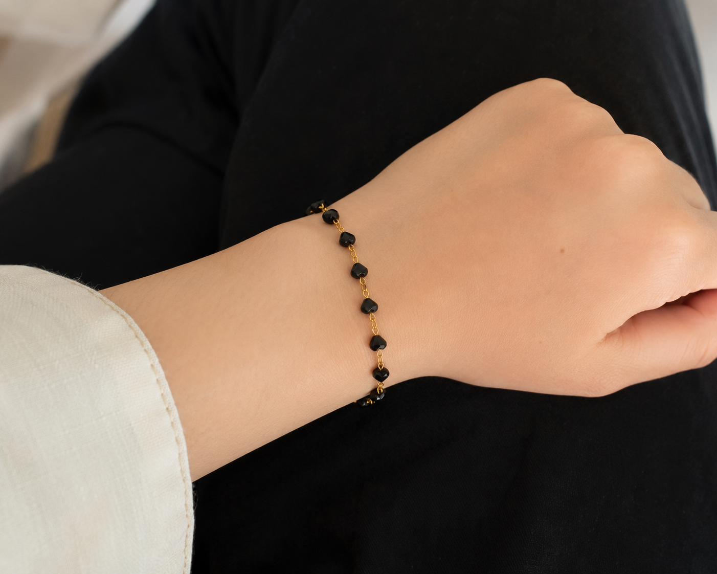 A delicate gold chain bracelet with small black beads worn on a person's wrist, shown against a dark black top and light beige skin tone.