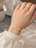 A close-up of a person's hand wearing a slim gold bangle bracelet with small clear stones, along with a matching gold ring, styled with a cream-colored blouse.