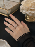 A close-up of a hand wearing a delicate silver ring with small sparkling stones, styled on a dark tabletop with a decorative glass box and cream flowers in the background.
