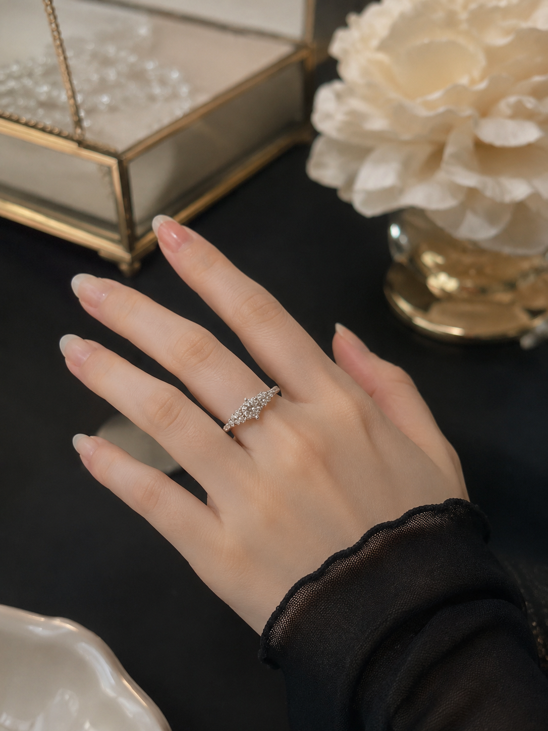 A close-up of a hand wearing a delicate silver ring with small sparkling stones, styled on a dark tabletop with a decorative glass box and cream flowers in the background.