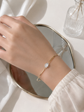 A delicate gold bangle bracelet with a single clear crystal setting worn on a person's wrist, shown against a soft white background with a mirror and glass bottle.