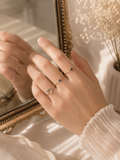 A close-up of a hand wearing delicate gold rings, including one with a small blue stone and another with a circular clear gemstone, posed in front of a decorative gold mirror with dried flowers in the background.