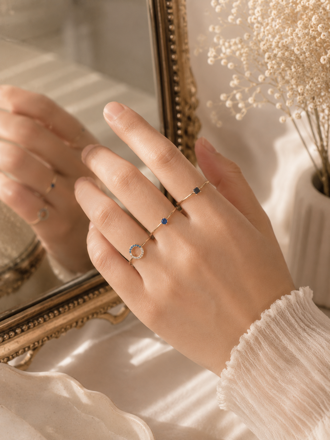 A close-up of a hand wearing delicate gold rings, including one with a small blue stone and another with a circular clear gemstone, posed in front of a decorative gold mirror with dried flowers in the background.