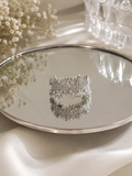 A wide silver-toned cuff bracelet covered in sparkling clear crystals, displayed on a round mirrored tray with soft cream fabric and dried flowers in the background.