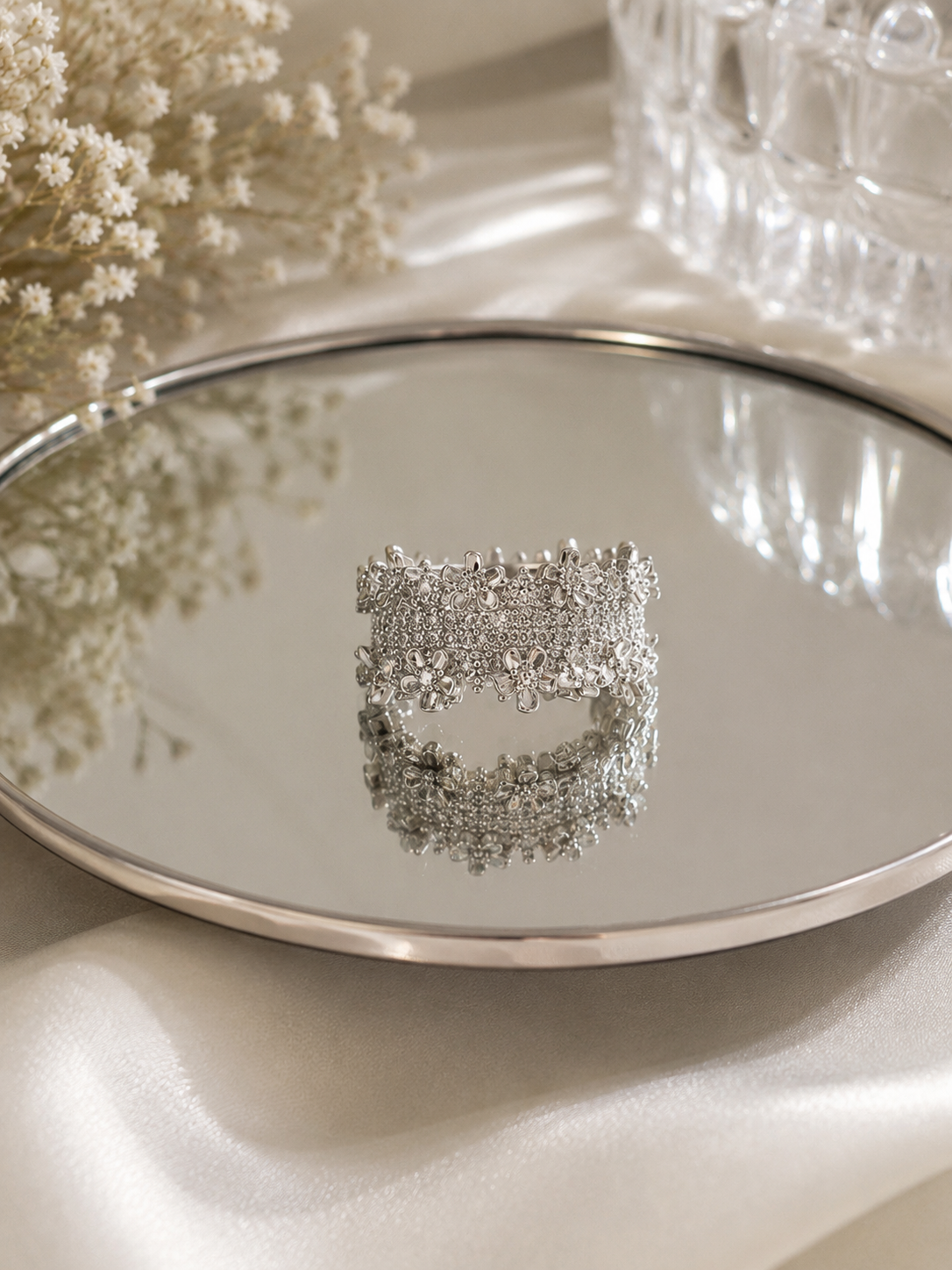 A wide silver-toned cuff bracelet covered in sparkling clear crystals, displayed on a round mirrored tray with soft cream fabric and dried flowers in the background.