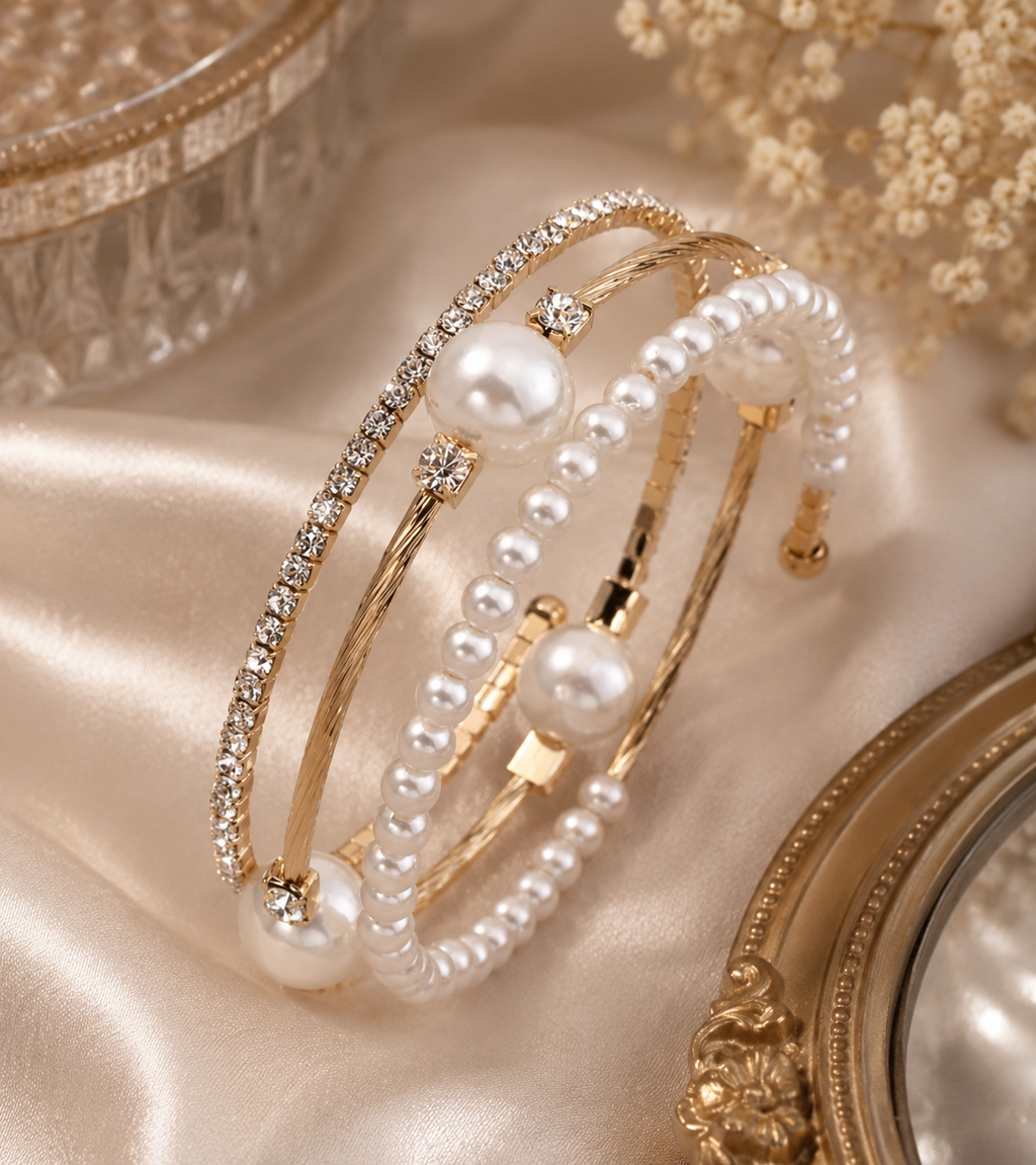 Three elegant gold-toned bangles styled with white faux pearls and clear rhinestones, arranged on a soft champagne satin fabric with decorative vintage mirror frames.