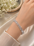 A close-up of a person's wrist wearing a sparkling silver-toned bracelet adorned with clear crystals, set against a soft beige satin background with white baby's breath flowers in a decorative bowl.
