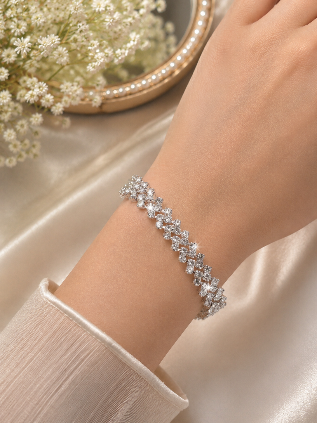 A close-up of a person's wrist wearing a sparkling silver-toned bracelet adorned with clear crystals, set against a soft beige satin background with white baby's breath flowers in a decorative bowl.