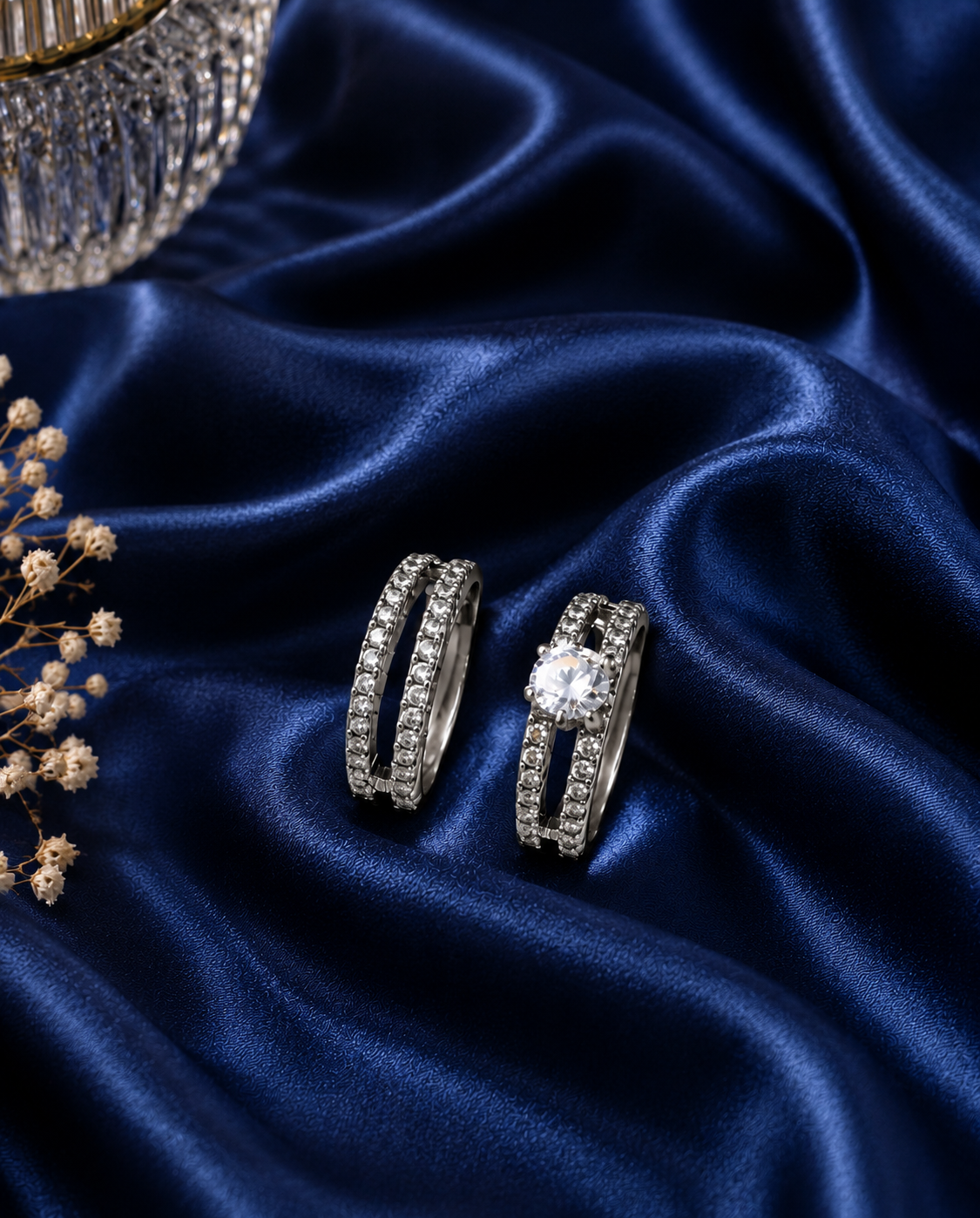 A pair of silver-toned rings displayed on draped dark blue satin fabric, with one ring featuring a large round clear center stone and the other showing a channel-set row of small clear stones. A partial crystal object and dried floral stems appear at the edges as styling props.
