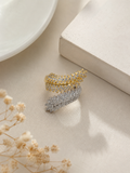 Two feather-shaped rings, one gold and one silver, both paved with clear stones, displayed on a textured beige fabric surface with neutral props.