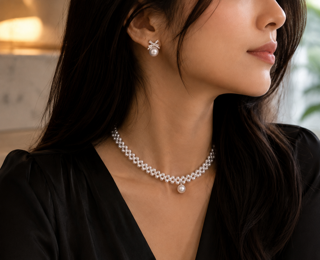 A close-up lifestyle shot of a woman wearing a delicate pearl necklace and matching pearl stud earring. The necklace features a silver-toned trellis base holding multiple white pearls, with a larger central pearl hanging at the center front.