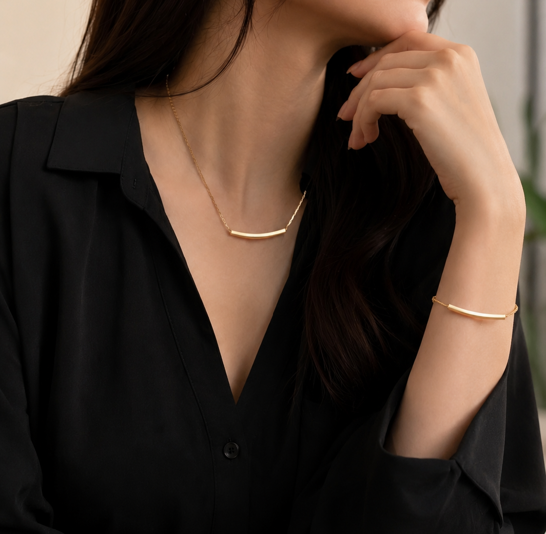 A close-up lifestyle shot of a woman wearing a delicate gold necklace and a matching gold bangle bracelet, styled with a black button-down shirt.