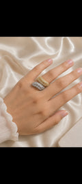 A close-up of a hand wearing three stacked textured rings in gold, silver, and white metals, resting on draped cream satin fabric.