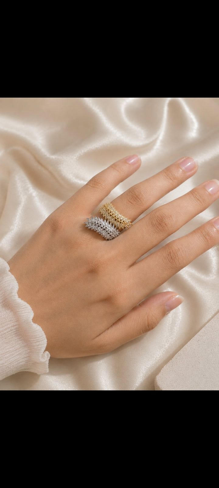 A close-up of a hand wearing three stacked textured rings in gold, silver, and white metals, resting on draped cream satin fabric.